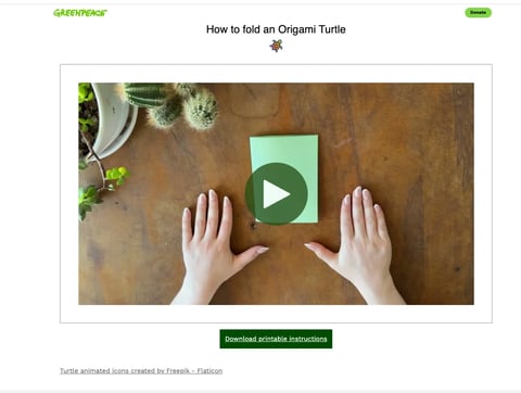 Overhead view of hands positioning a green paper square on a wooden surface with a play button overlay